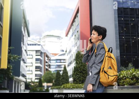 A businesslike modern woman stands on the street of the business center and talks on the phone. The woman is dressed like a hipster. A middle-aged wom Stock Photo