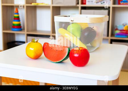 Orange watermelon slice and red apple stand on a white table with a basket of toy produce in a colorful playroom inviting pretend grocery play Stock Photo