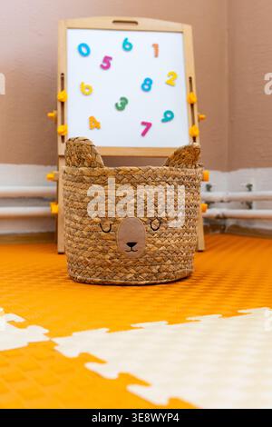 A woven storage basket with a sleepy bear face sits on a bright foam mat while a small number board rests behind. Cozy storage adds charm Stock Photo