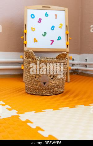Woven bear face basket placed on bright interlocking foam tiles with a whiteboard of colorful numbers in the background. Friendly storage brings warmt Stock Photo
