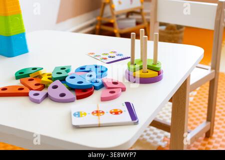 A white table holds a colorful wooden shape sorter with pegs and pattern cards in a sunny classroom. Open ended play supports matching colors sorting Stock Photo