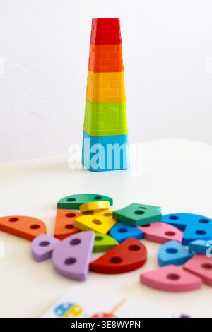 A tall rainbow stacking tower rises on a white table while chunky wooden pieces scatter nearby. Bold colors invite toddlers to sort stack and imagine Stock Photo