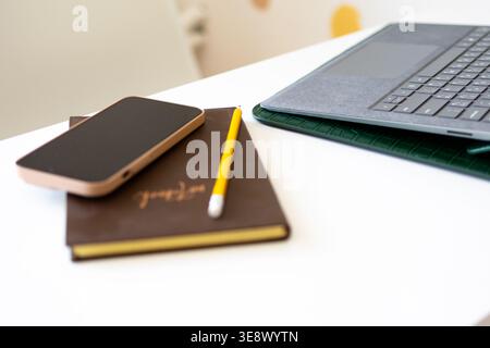 Smartphone and yellow pencil lie on a brown notebook next to a slim laptop on a white desk. Clean lines and soft light create a modern study space Stock Photo