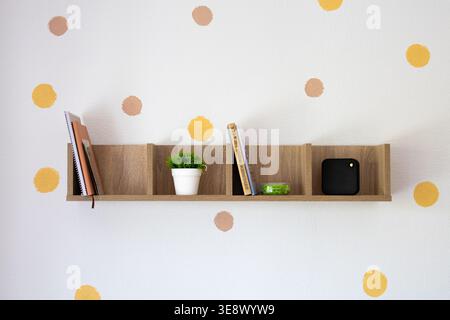 Floating wall shelf on a dotted backdrop holding books a potted plant a small gadget and tape. Simple arrangement and clean lines create stylish Stock Photo