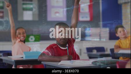 Boy in red shirt raising hand and holding pencil in elementary classroom with digital overlays Stock Photo