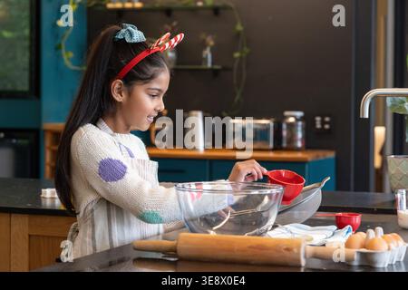 Asian female child in polka-dot sweater and striped apron, measuring with red cup over glass bowl Stock Photo