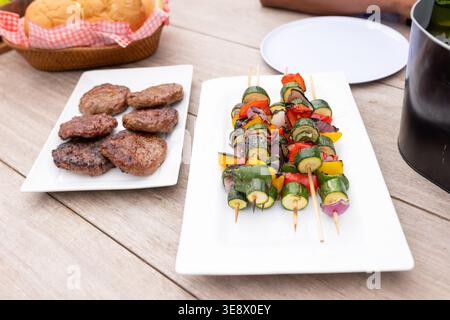 Vegetable skewers on wooden sticks resting on white platter at patio with burger patties Stock Photo