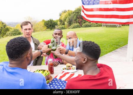 Diverse male friends sitting at table in yard by American flag, clinking bottles, eating burgers Stock Photo