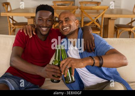 Two African American men on sofa clinking green beer bottles toasting, showing ring and bracelet Stock Photo