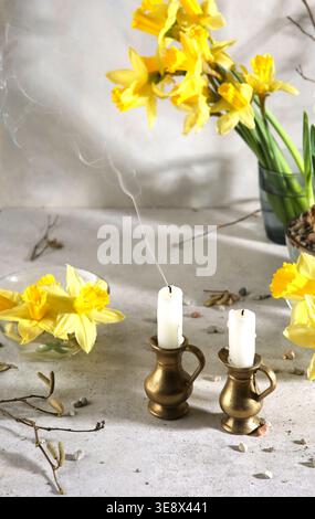 Two candles with daffodil flowers indoor. Extinguished candles in jug candlesticks. Stock Photo