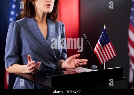 Medium shot of middle aged Caucasian woman speaking at podium beside United States flag, gesturing during public address. Useful for political campaign, leadership Stock Photo