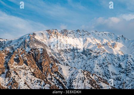 A stunning view of a snow-covered mountain range, showcasing rugged peaks and rocky terrain against a clear blue sky with wispy clouds. Stock Photo