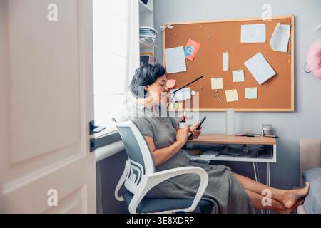 middle-aged Asian woman with short black hair sits at a desk, using a smartphone. A bulletin board with notes and papers is in the background. Stock Photo
