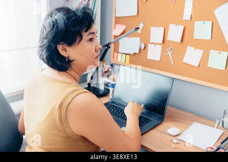 young Asian woman with short black hair works on a laptop at a wooden desk. A corkboard with notes and stationery is in the background. Stock Photo