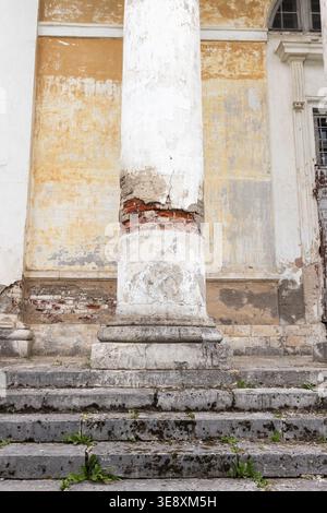 A weathered classical column stands on mossy stone steps in front of a historic building with peeling plaster, exposed brick and a crumbling facade, e Stock Photo