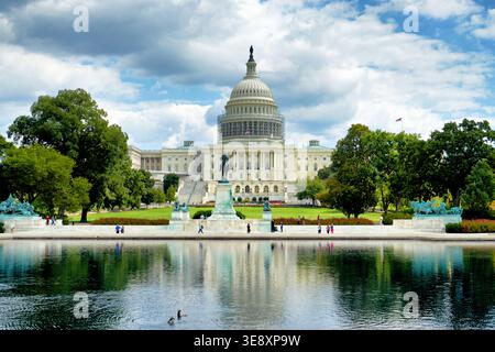 Visitors walk around the reflecting pool near the Capitol building in Washington DC. The sun shines over the scene with clouds in the sky. Stock Photo