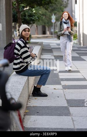 Two women in 20s sitting and walking along campus walkway holding notebooks and phone, copy space Stock Photo