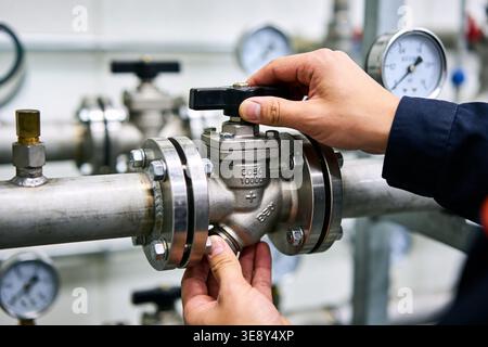 A worker's hands are carefully turning a valve on a complex network of metal pipes and pressure gauges in an industrial setting Stock Photo