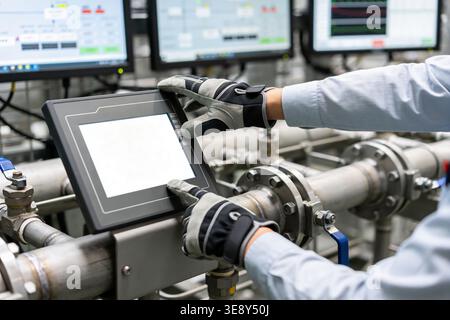 A person wearing work gloves interacts with a digital control panel in a factory setting with pipes and monitors visible Stock Photo