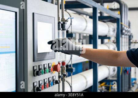 A person wearing protective gloves interacts with a digital interface on a complex industrial control system, surrounded by pipes and machinery Stock Photo
