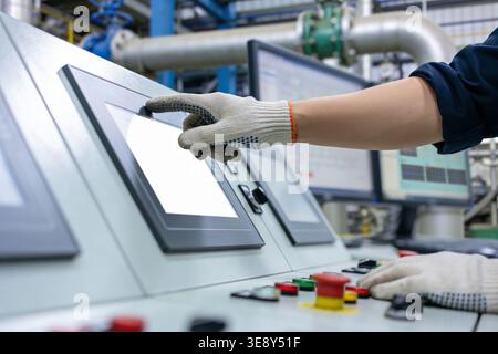 A worker wearing protective gloves touches a blank screen on a sophisticated control system in a factory setting, with monitors displaying data in the Stock Photo