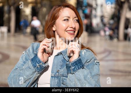 Red haired woman walking in a city street, cheerfully smiling and holding headphones, enjoying music and sunny weather Stock Photo