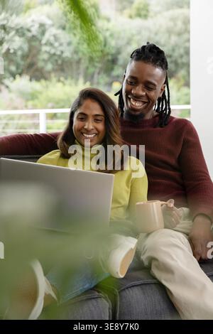 South Asian woman and African American man couple sitting on grey couch on porch, using laptop. Sofa, duo, device, home, lifestyle, cozy, natural Stock Photo