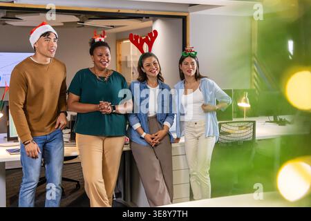 Diverse coworkers standing by desks in open plan office, wearing business casual and holiday hats. Team, celebration, festive, colleagues, workstation, monitors, lights Stock Photo
