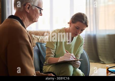 Senior Caucasian man consulting young adult caregiver in nursing home, discussing health needs while caregiver taking notes Stock Photo
