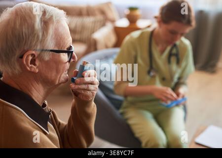 Senior man using inhaler during respiratory care consultation in nursing home, nurse preparing medication nearby Stock Photo