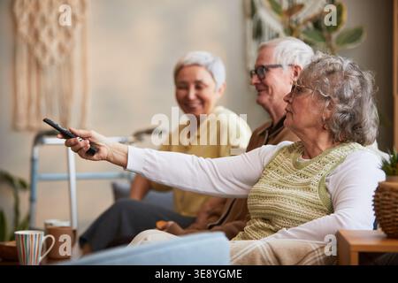 Senior Caucasian woman using remote control while sitting with senior friends in nursing home, representing assisted living leisure Stock Photo