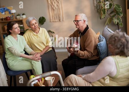 Senior man playing guitar for senior women and young adult caregiver in nursing home, supporting social engagement Stock Photo