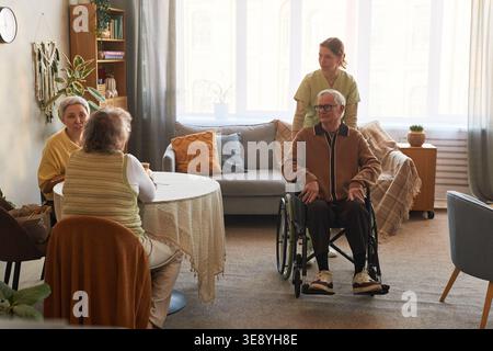 Senior man with disability sitting in wheelchair while caregiver assisting and senior women talking in nursing home Stock Photo