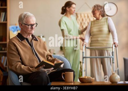 Senior Caucasian man reading book in nursing home while caregiver assisting senior woman with walker, showing elder care support Stock Photo