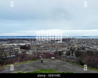 Wide elevated view over Edinburgh showing historic buildings, distant Firth of Forth and two visitors on a hilltop. Stock Photo
