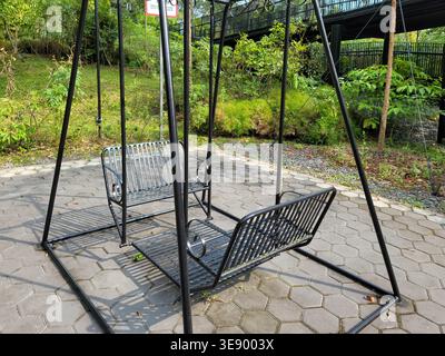 Black metal swing sits on paved stone ground. Garden greenery surrounds the swing's base. Sunlight casts shadows across the swing frame. Metal seats a Stock Photo