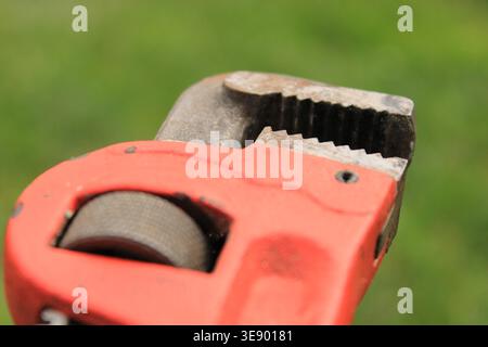 A red hand tool is shown in close detail. The tool has a gripping mechanism made for various plumbing tasks. It rests on green grass, highlighting its Stock Photo