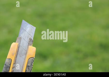 A person holds a utility knife with a yellow handle in front of a green grass background. The knife is open and ready for use in various tasks. Stock Photo