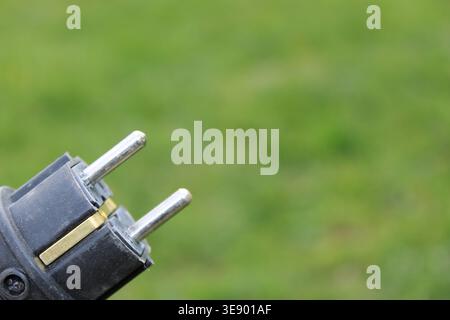An electric plug with two metal prongs is shown in close detail. The plug is set against a green lawn while the sun is shining. The scene captures the Stock Photo