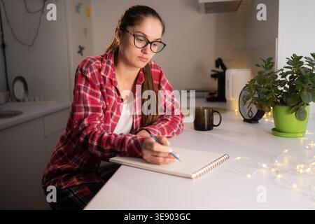 Young woman writing notes in paper notebook in cozy home kitchen. planning and housekeeping Stock Photo