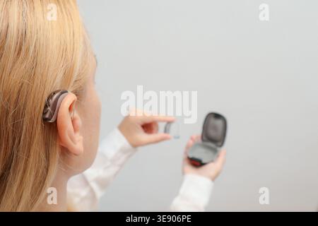 Blond woman applying Hearing Aid on ear at home. Deafness treatment, hearing solutions. Deaf woman wearing hearing aid. Digital hearing aid in woman's Stock Photo