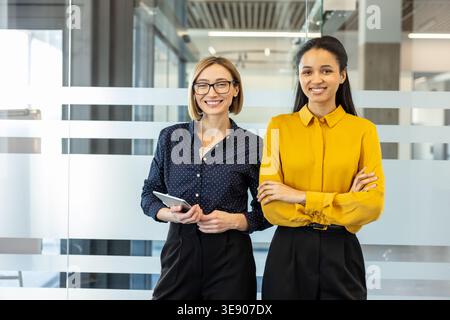 Two smiling professional business women, one holding a tablet and another with crossed arms, standing confidently in a bright contemporary office, illustrating professionalism, teamwork, and success Stock Photo