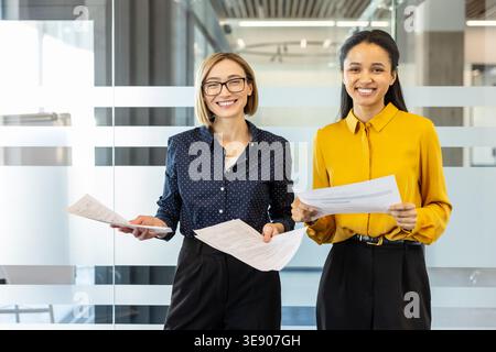 Two smiling multi-ethnic businesswomen are holding paperwork, standing in a brightly lit modern office demonstrating professional collaboration and teamwork Stock Photo