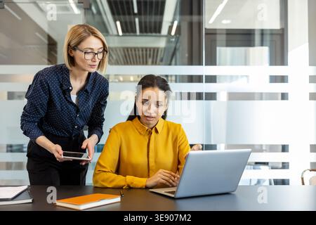 Two businesswomen are actively collaborating, discussing a business project and analyzing data on a laptop and digital tablet while working together in a modern, bright office environment Stock Photo