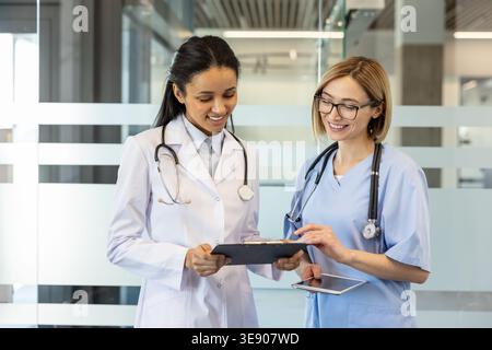Two smiling female doctors, one wearing a white lab coat and the other blue scrubs, studying patient medical records on a clipboard and digital tablet in a modern clinic corridor Stock Photo