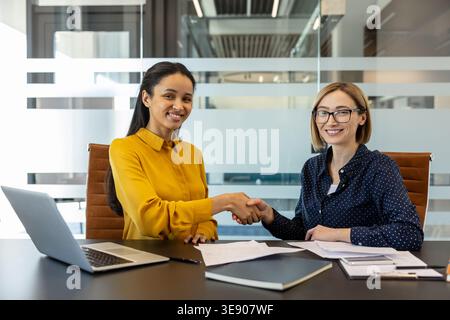 Two smiling multiethnic businesswomen seated at a modern office desk shaking hands after signing documents, celebrating a successful professional agreement and collaborative partnership Stock Photo