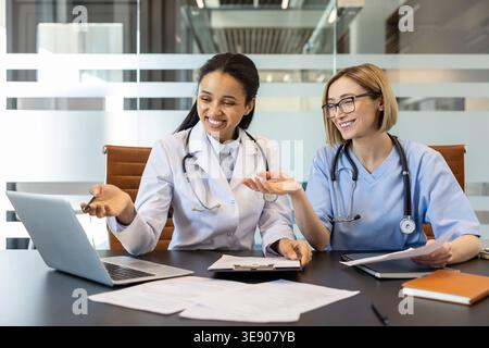 Two female medical professionals are smiling while collaborating on patient data. Discussing information shown on a laptop during an office meeting. Symbolizing teamwork and modern healthcare analysis Stock Photo