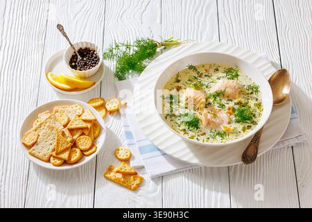 Swedish laxsoppa creamy salmon soup with chunks of fish and fresh dill in a bowl served with crackers, lemon slices and peppercorns on a white wooden Stock Photo