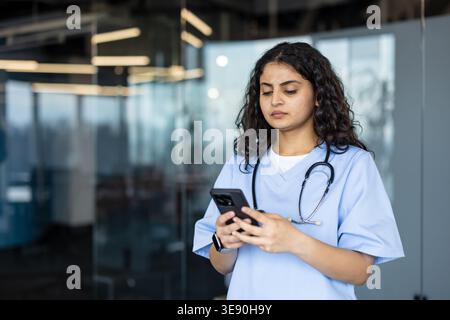 Young indian female doctor or nurse in light blue scrubs and stethoscope using a smartphone for telehealth, communication or medical app in a modern h Stock Photo