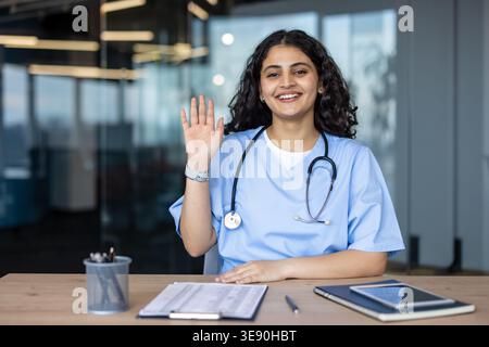 Young female healthcare professional wearing scrubs and stethoscope, sitting at a desk in a modern office, smiling, looking at the camera, and waving Stock Photo